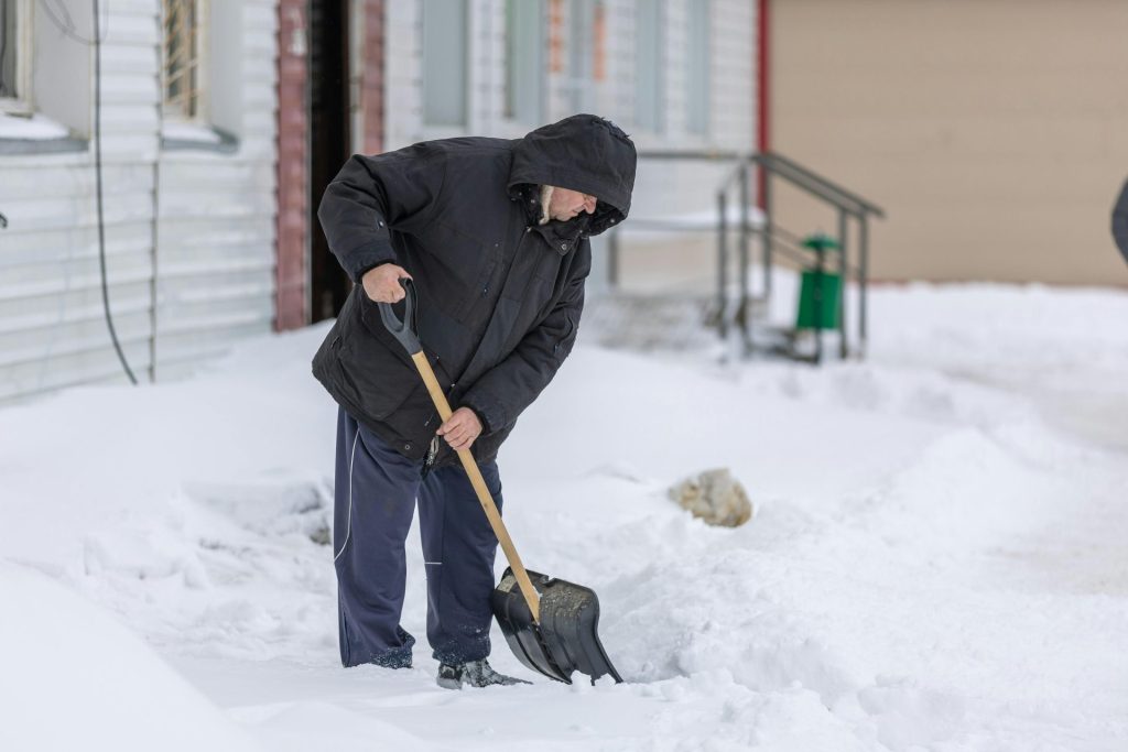 Ein erwachsener Mann schaufelt an einem kalten Wintertag Schnee und zeigt damit Aktivitäten bei winterlichen Bedingungen.